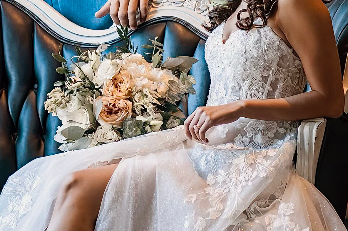 Bride in a white wedding dress holding a bouquet of flowers, sitting on a blue chair.