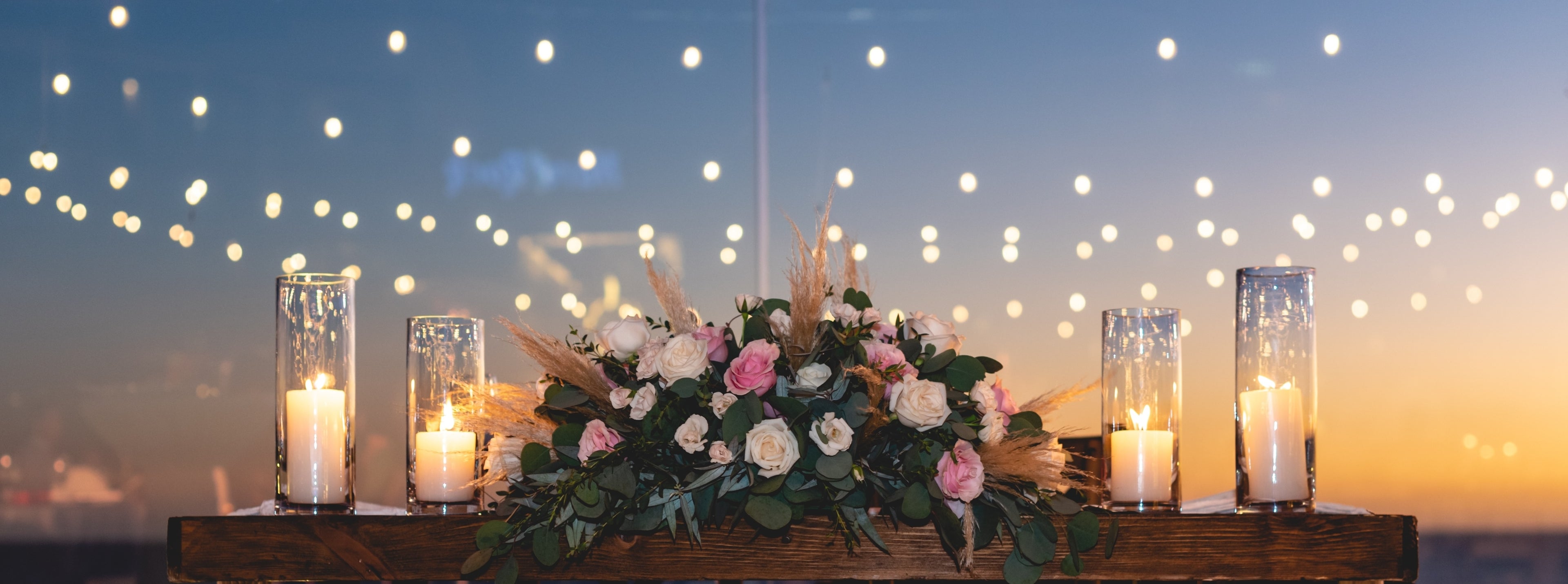 Decorative floral arrangement with candles on a wooden platform against a twilight sky with string lights.