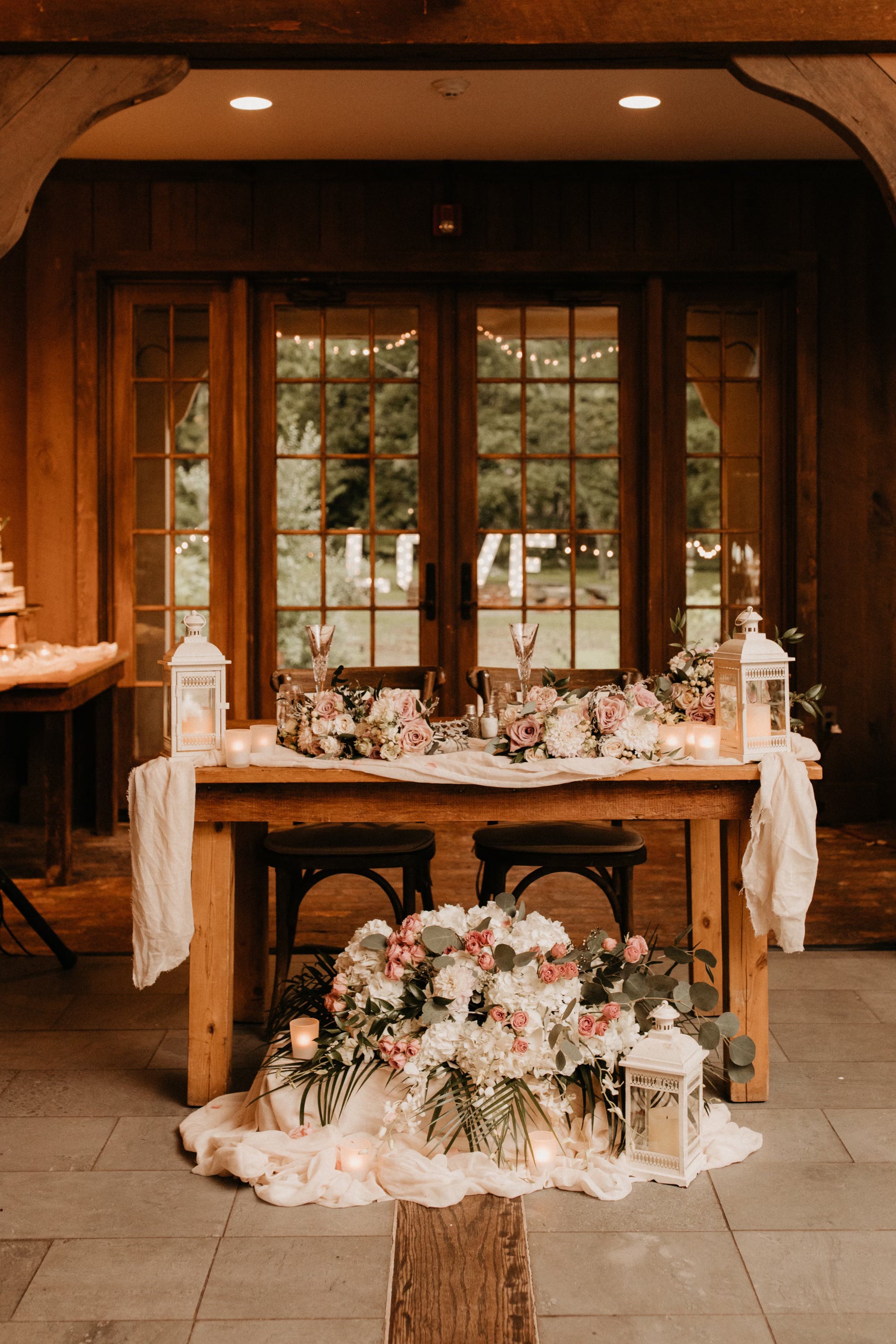 Decorated wooden table with floral arrangements and lanterns in a rustic setting.
